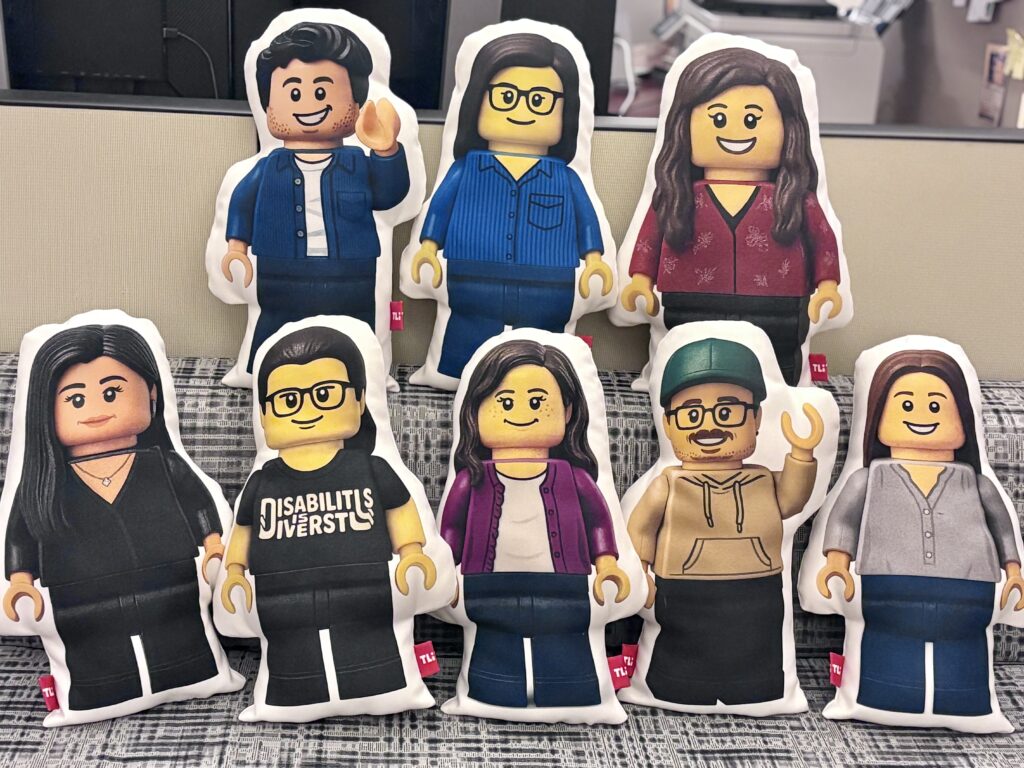 Eight pillow figures designed as LEGO-style characters are arranged in two rows on a patterned couch in the TLi office.